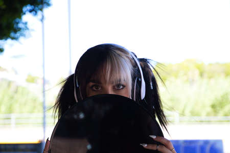 Young and beautiful girl with a punk and heavy tendency holds a vinyl record in her hands. She plays at hiding behind the vinyl by making different gestures. Retro and 80's conceptの写真素材