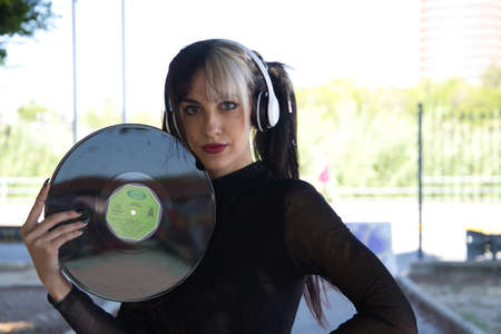 Young and beautiful girl with a punk and heavy tendency holds a vinyl record in her hands. She plays at hiding behind the vinyl by making different gestures. Retro and 80's conceptの写真素材