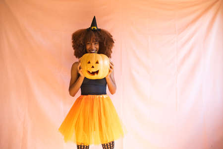 young african american woman in witch costume for halloween party. She is wearing a witch hat, orange skirt and black angel wings. The woman smiles at the camera while playing with the pumpkin.の写真素材