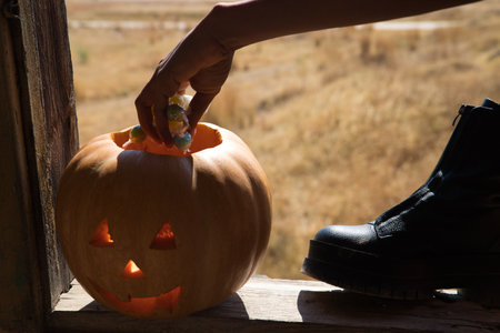 hand detail of African American woman picking up and putting candy into a Halloween pumpkin. The woman is sitting in the window frame of an abandoned building.の写真素材
