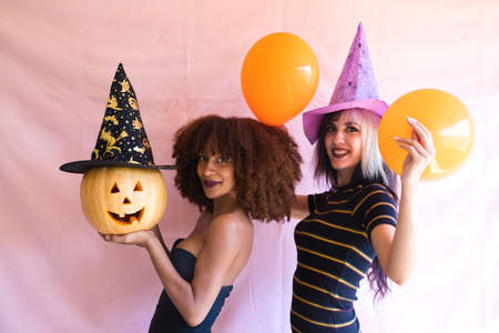 two beautiful young women, one blonde and the other African-American, dressed as witches and holding a pumpkin, are having fun at a Halloween party. The women are happy.の写真素材