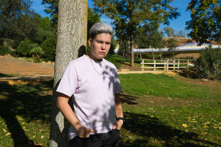 gender non-binary and androgynous person doing different postures and leaning on a tree. He is looking at the camera. Concept of non-binary and androgynous. Concept of diversity and expressions.の写真素材