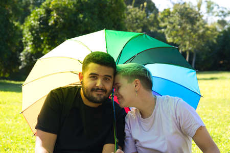 non binary gender person and a gay man are sitting in the park, covering themselves from the sun with a gay pride umbrella. they are posing for the camera and very happy. Diversity and gay pride.の写真素材