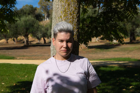 gender non-binary and androgynous person doing different postures and leaning on a tree. He is looking at the camera. Concept of non-binary and androgynous. Concept of diversity and expressions.の写真素材
