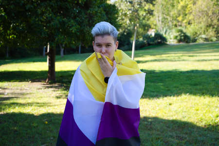 gender non-binary person is holding the non-binary pride flag on his shoulders while kissing it. He is looking at the camera. Concept of non-binary and androgynous. Diversityの写真素材