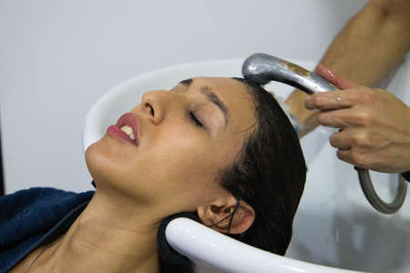 Transgender woman lying down having her head hair washed in beauty salon by professional hairdresser. Beautiful client feeling relaxed as hairdresser massages head in beauty salon hair salon.の写真素材