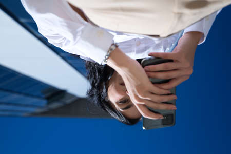 young, beautiful woman is consulting her mobile phone at the entrance door of the building where she works. The photo is taken from below and the building can be seen. Concept businesswoman.の写真素材