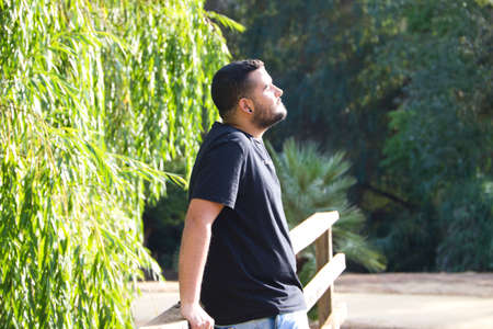 A gay man of Latin origin, dark and bearded, stands pensively looking nowhere in the park. He is sad, dazed and depressed. Concept of expressions of sadness and depression.の写真素材