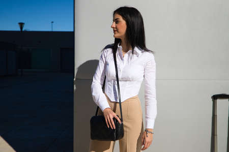 young, beautiful woman is standing at the entrance of the building where she works. The woman is wearing brown trousers and a white shirt with a black handbag. Concept of a working woman.の写真素材