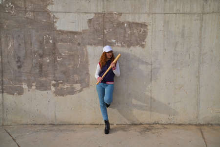 young and beautiful redhead woman is happy with baseball cap, jacket, baseball bat and jeans, she is posing in front of gray concrete background. Concept sports and recreations.の写真素材