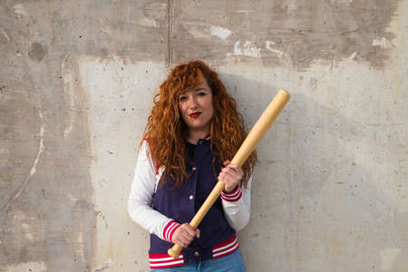 young and beautiful redhead woman is happy with jacket, baseball bat and jeans, she is posing in front of gray concrete background making different expressions. Concept sports and recreations.の写真素材