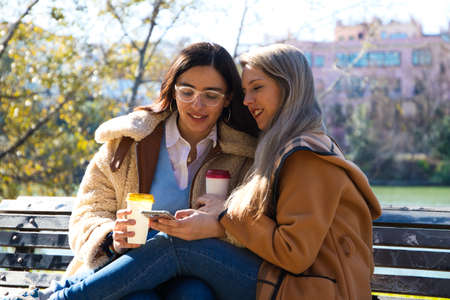 A lesbian couple sitting on a bench checking their social networks over a cup of coffee. The women are young and recently married. Concept lgtb, gays and lesbians. Rights and equalityの写真素材