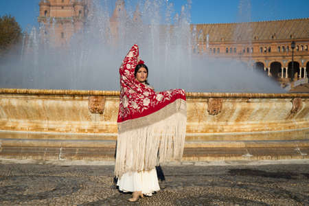 Flamenco dancer woman, brunette and beautiful typical spanish dancer is dancing with a red manila shawl in front of a fountain in a square in Seville. Flamenco concept of cultural heritage of humanityの写真素材
