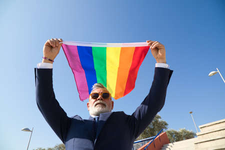 Mature gay man, executive, grey-haired, bearded, sunglasses, jacket and tie, waving the new lgbtiq+ pride flag in the wind under a blue sky. Mature gay man concept, lgbt, pride, equality, rights.の写真素材