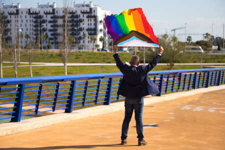 Mature gay man, executive, gray hair, beard, sunglasses, jacket and tie, waving the new gay pride flag in the wind, photo taken from behind. Concept of mature gay man, lgtb pride, equality, rights.の写真素材