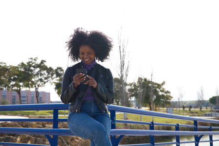 Young, beautiful Afro-American woman with mobile phone shopping online, checking her email and social networks in the park. The woman is wearing casual and comfortable clothes and is happy.の写真素材