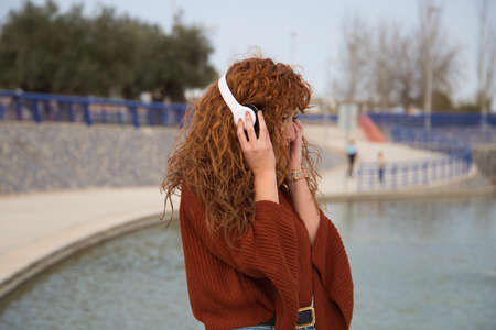 Young and beautiful red-haired woman listening to music with white headphones in a park where there is a big lake. The woman is happy. Concept expressions and happiness, music and dance.の写真素材