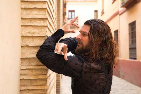 Long haired man dancing flamenco with black shirt and red roses. He makes dancing postures with his hands in a typical narrow street of Seville. Flamenco dance concept cultural heritage of humanity.の写真素材