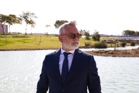 Handsome mature man, executive, gray hair, beard, sunglasses, jacket and tie, is in a park with an artificial lake posing for photo. Concept of mature man, grandfather, executive.の写真素材