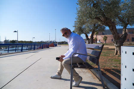 Handsome mature man with beard and gray hair is sitting thoughtfully on a park bench while consulting his mobile phone. The man is dressed in beige trousers, white shirt. Concept of old ages.の写真素材