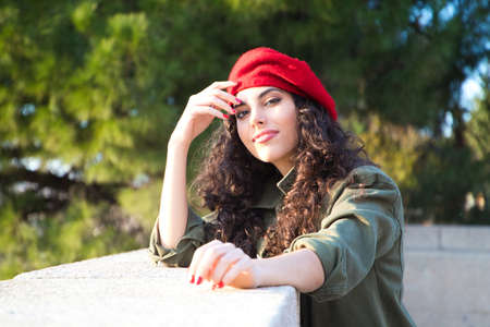 Beautiful young latin woman with curly brown hair wearing a red cap and dressed in casual clothes is sightseeing in Europe. She is posing for photos. Mediterranean pine tree in the background.Tourismの写真素材
