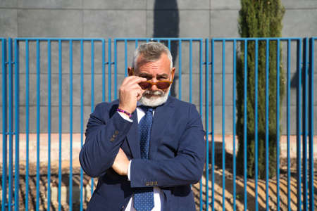 Mature, grey-haired, bearded man with sunglasses, jacket and tie makes different expressions for the photo, looks over his sunglasses and touches his lips. Concept various expressions.の写真素材