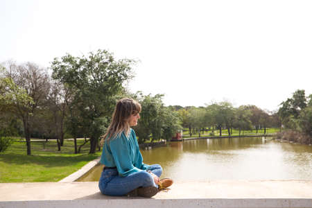 Pretty, blonde, young woman is sitting on the bridge over the river while looking at the horizon smiling. the sky is white and leaden. Concept of holidays and free time. Various expressions.の写真素材