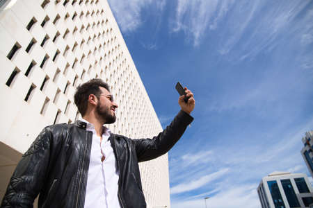 handsome young man with beard, sculptural body is taking a selfie with his mobile phone in the street.の写真素材