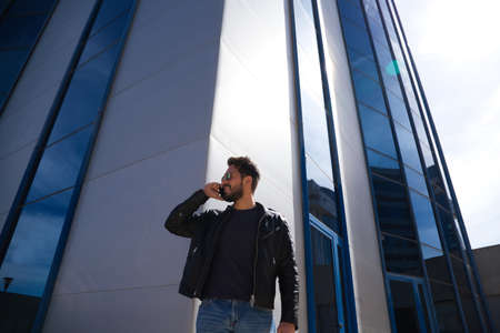 Handsome young man with beard, sculpted body and sunglasses is talking on the phone in front of a glass building. The man is wearing jeans and a leather jacket.の写真素材