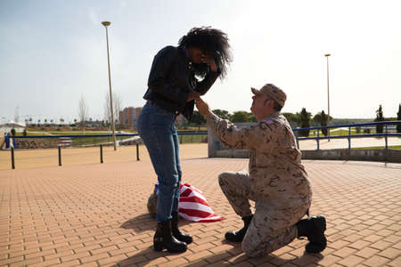 American soldier on his knees asking for marriage by holding the hands of his partner an Afro-American woman and giving an engagement ring. Concept patriotism, war, soldier.の写真素材