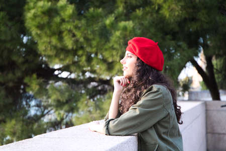 Beautiful young latin woman with curly brown hair wearing a red cap and dressed in casual clothes is sightseeing in Europe. She is looking at the horizon. Mediterranean pine tree in the background.の写真素材