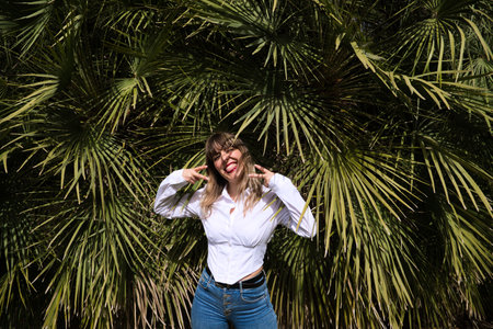 Pretty, blonde, young woman having fun making different expressions in front of green palm trees in the park. Holiday and leisure time concept. Various expressions.の写真素材
