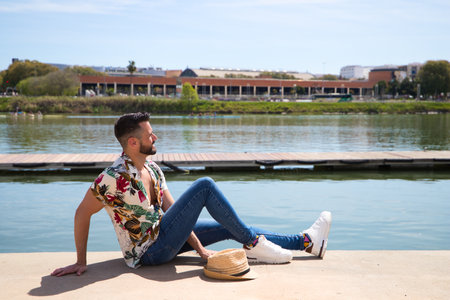 Handsome young man is wearing a shirt with Hawaiian flowers, a hat and is sitting on the river bank at the jetty sunbathing. Holiday and leisure time and travel concept.の写真素材