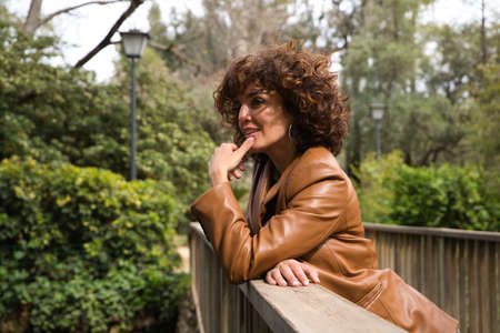 beautiful woman with curly hair leaning on the railing of a wooden bridge in a park with abundant vegetation The woman is dressed in modern and trendy clothes. Travel and holidays concept.の写真素材