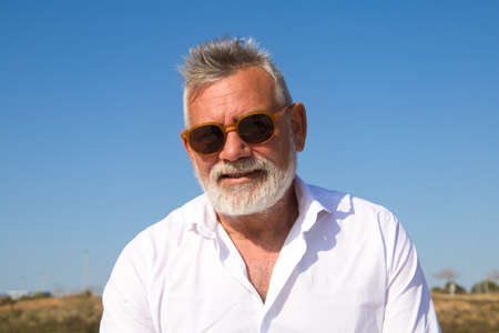 Handsome mature man with beard and gray hair is sitting pensively on a park bench while looking at the camera. The man is dressed in beige trousers, white shirt. Concept of old age and technology.の写真素材