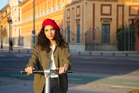 Beautiful young latin woman with curly brown hair wearing red cap and dressed in casual clothes is sightseeing in Europe visiting the city on scooter. Tourism concept.の写真素材