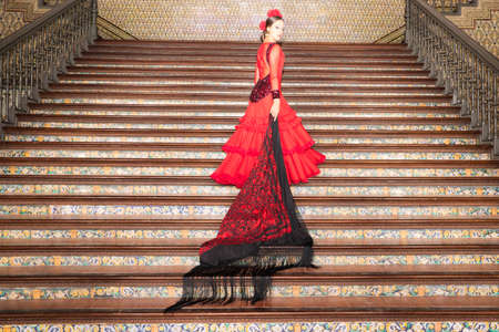 A beautiful teenage woman dancing flamenco goes up the stairs clutching a fringed and embroidered shawl in her hand. She is wearing a red dress with a frill. Flemish cultural heritage of humanity.の写真素材