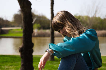 Young, pretty blonde woman dressed in jeans and boots is sitting at a wooden table in the park. In the background you can see the river on a sunny day. The woman is happy. Concept holidays and free.の写真素材