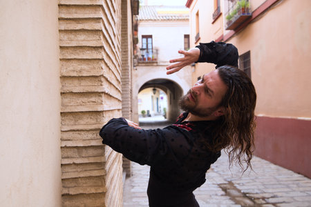 Long haired man dancing flamenco with black shirt and red roses. He makes dancing postures with his hands in a typical narrow street of Seville. Flamenco dance concept cultural heritage of humanity.の写真素材