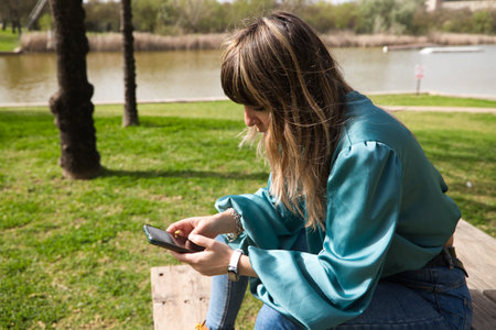 A young, pretty, blonde woman is sitting at a wooden table in the park, looking at her mobile phone and smoking a cigarette. In the background you can see the river on a sunny day. stop smokingの写真素材