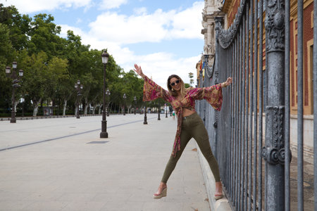 Young, blonde and beautiful woman is on holiday visiting Seville. The woman is holding on to the fence of a monument while having fun. Holiday and tourism concept.の写真素材