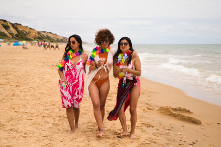 three young and beautiful latin women with a glass of blue cocktail stroll along the beach happily talking to each other and having fun. Holiday and travel concept.の写真素材