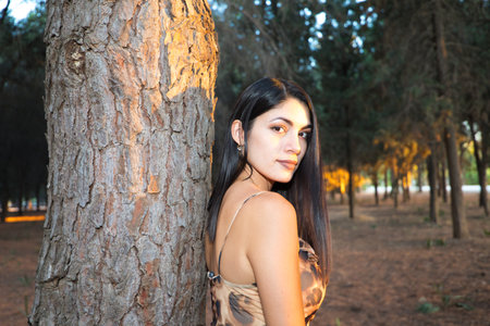 beautiful young woman from south america leaning on a tree in the forest at the golden hour. Rays of sunlight enter through the trees in autumn and create golden atmosphere.の写真素材