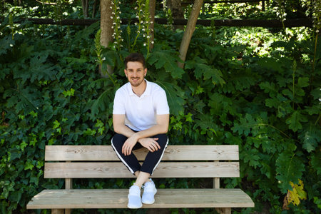 Handsome young man in white T-shirt and black trousers sitting on the wooden bench in the park waiting impatiently for someone to arrive. The man makes different body expressions. Concept expression.の写真素材