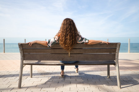 Blonde woman with curly hair sitting on a bench looking at the horizon of the sea. The woman is relaxed and nostalgic looking at the sea. Concept relaxation and peace.の写真素材