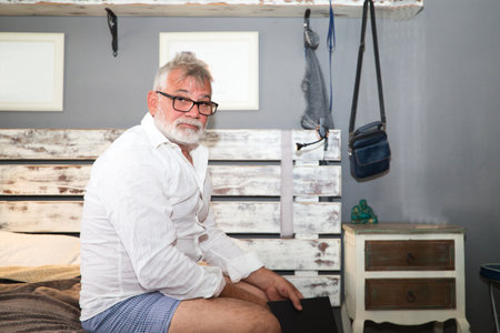 Senior man with white beard sitting on bed with a book in his hands. Man is in pajamas and has glasses so he can read. Seniors and grandparents concept.の写真素材