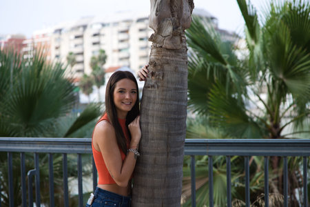 Young and beautiful woman leaning on the trunk of a palm tree. Woman is happy and relaxed and enjoying the sunny day in Seville, Spain. The woman is wearing jeans and orange top.の写真素材