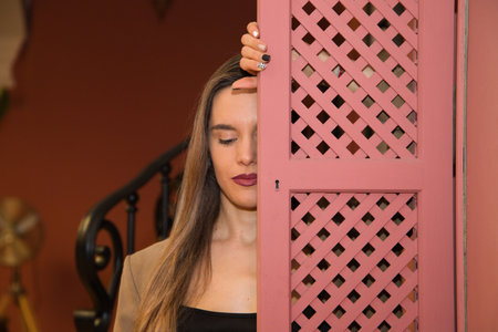 Beautiful young woman behind a pink wooden window cover. The woman is sad and looks at the ground with sorrow and depression.の写真素材