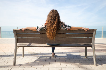 Blonde woman with curly hair sitting on a bench looking at the horizon of the sea. The woman is relaxed and nostalgic looking at the sea. Concept relaxation and peace.の写真素材