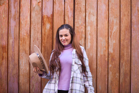 Young and beautiful Spanish woman from Seville with brown hair and plaid shirt holds a hat in her hand and offers it to the camera. The woman is happy and in the background a wooden door.の写真素材